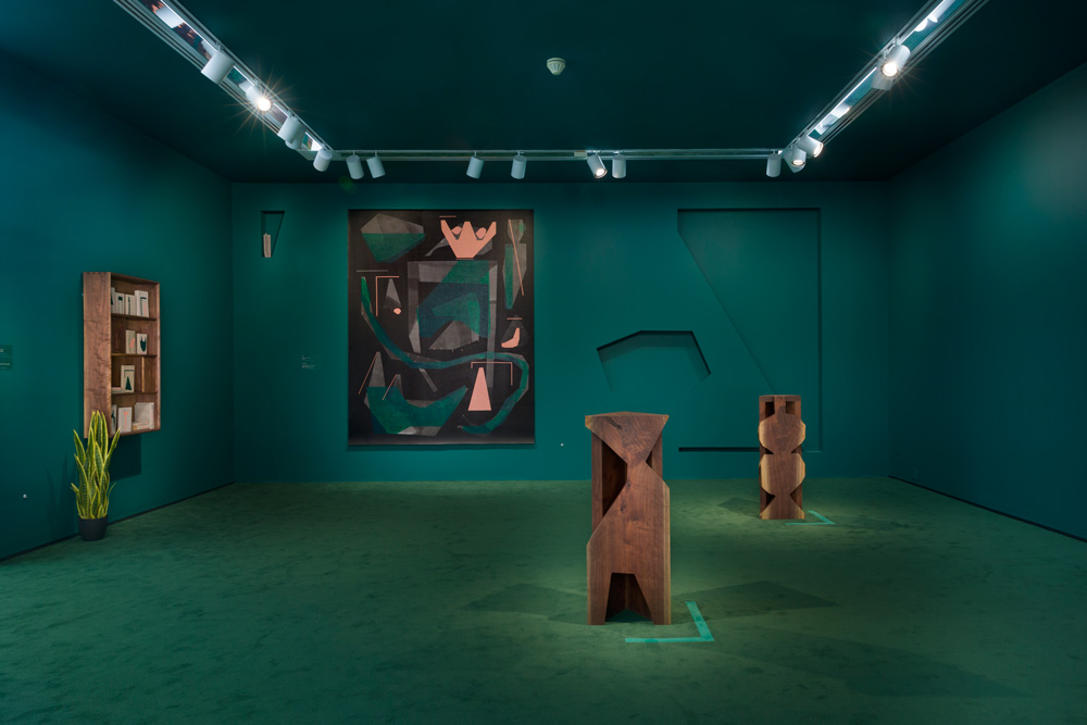 Photo of gallery with forest green-colored walls, two pedestal-shaped sculptures, and an abstract painting on the wall