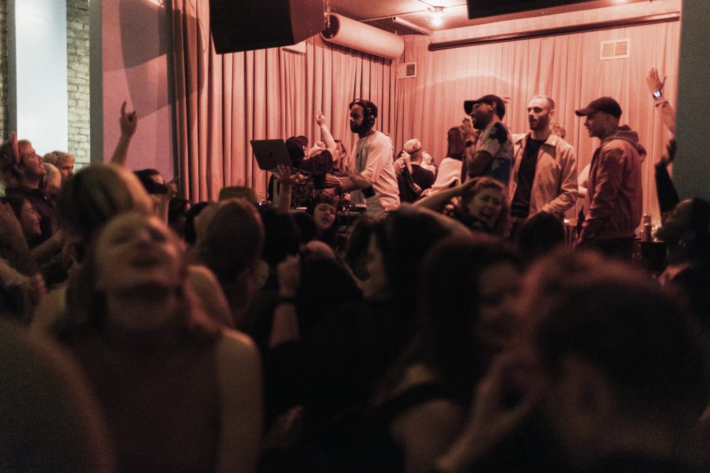 Crowd dancing in front of a DJ on a stage with a laptop, some onlookers and pink curtains behind him.