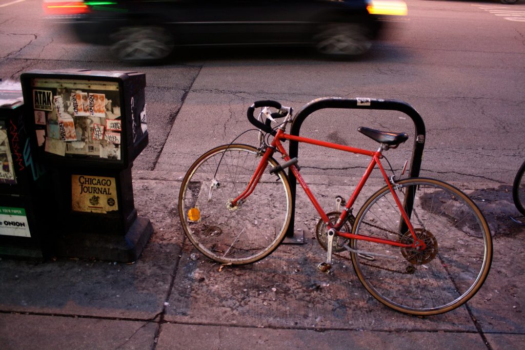Photo of red bike attached to bike rack on sidewalk next to Chicago Journal newsbox with stickers covering front.