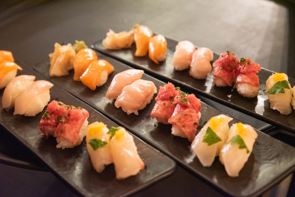 Array of sushi in yellow and and pink hues laid out on three dark rectangular trays.