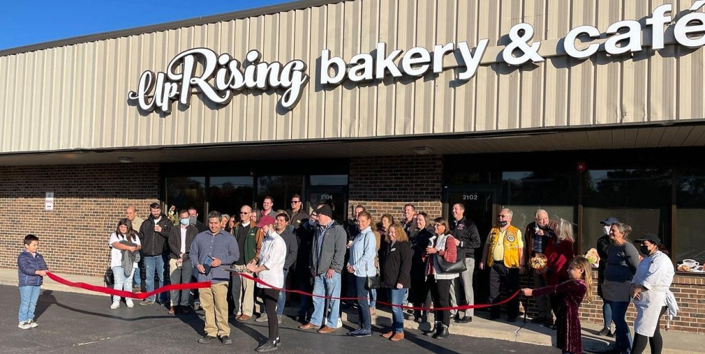 Photograph of crowd of people standing in front of a storefront with words "Uprising Bakery & Cafe" with red ribbon being cut.