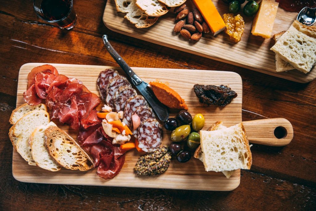 Photograph above a wooden cutting board with a display of charcuterie, slices of bread, olives and mustard.