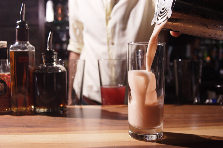 Bartender pouring out a fizzy pink cocktail into a tall glass on a wood bar.