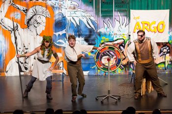 Three performers in various costumers next to a spinning price wheel in front of a banner that reads Argo with a mural of a Roman soldier.
