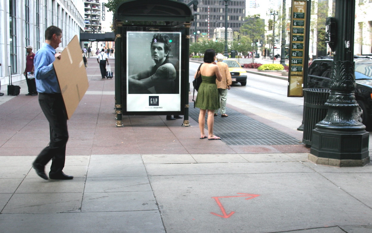 A photo of people waiting at a bus stop on State Street. One man holds a flattened cardboard box.