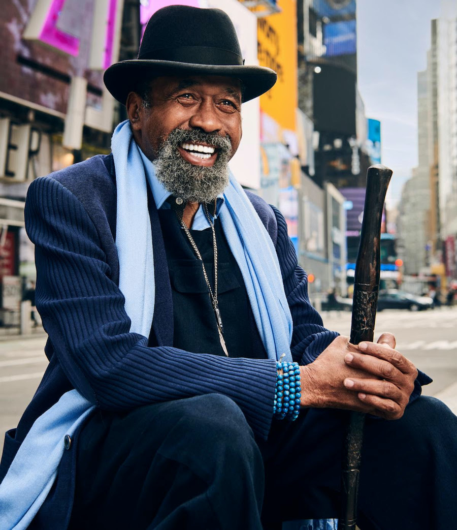 Ben Vereen smiling while wearing a suit jacket, scarf and hat, sitting with a city backdrop behind him.
